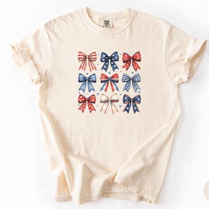 May include: An ivory short-sleeve t-shirt featuring a central graphic print of nine decorative bows arranged in a grid. The bows display various red, white, and blue patterns, including stripes, stars, and solid colors, accented by small blue stars.