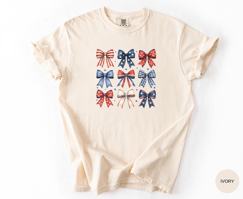 May include: An ivory short-sleeve t-shirt featuring a central graphic print of nine decorative bows arranged in a grid. The bows display various red, white, and blue patterns, including stripes, stars, and solid colors, accented by small blue stars.