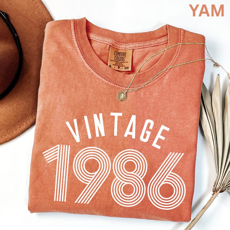 May include: A coral-colored t-shirt with the text "VINTAGE 1986" in white. The shirt has a crew neck and a small tag. A gold necklace with a pendant and a brown hat are also visible.