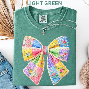 May include: Light green t-shirt featuring a large, colourful patchwork bow design. The bow incorporates floral, polka dot, and gingham patterns in pink, yellow, blue, and purple. A silver necklace with a pendant is draped on the shirt. The text "LIGHT GREEN" is at the top.