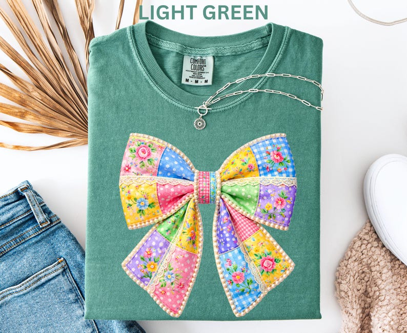 May include: Light green t-shirt featuring a large, colourful patchwork bow design. The bow incorporates floral, polka dot, and gingham patterns in pink, yellow, blue, and purple. A silver necklace with a pendant is draped on the shirt. The text "LIGHT GREEN" is at the top.