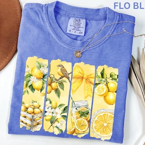 May include: A blue Comfort Colors t-shirt with a lemon-themed graphic. The graphic features yellow panels with lemons, a bird, a bow, and a jar of lemonade. The shirt has a crew neck and short sleeves. The text "FLO BLUE" is at the top.