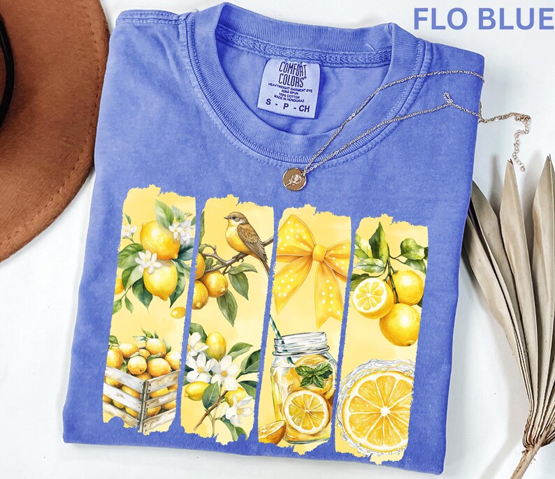 May include: A blue Comfort Colors t-shirt with a lemon-themed graphic. The graphic features yellow panels with lemons, a bird, a bow, and a jar of lemonade. The shirt has a crew neck and short sleeves. The text "FLO BLUE" is at the top.