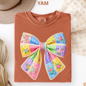 May include: A terracotta-coloured t-shirt with a large, colourful bow design. The bow is made of patchwork squares in various colours and patterns, including floral prints and polka dots. The shirt has a crew neck and the text "YAM" is visible.
