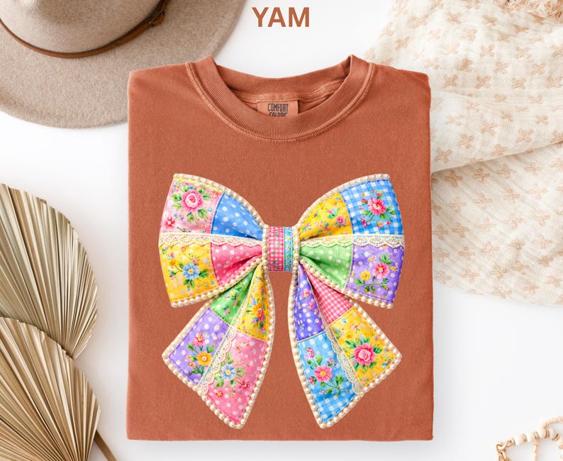 May include: A terracotta-coloured t-shirt with a large, colourful bow design. The bow is made of patchwork squares in various colours and patterns, including floral prints and polka dots. The shirt has a crew neck and the text "YAM" is visible.