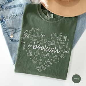 May include: A moss green crewneck t-shirt featuring a white graphic print of the word bookish in cursive script, surrounded by small line art illustrations of pumpkins, books, mugs, leaves, mittens, beanies, and bows in a heart shape.