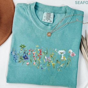 May include: Seafoam green t-shirt with a floral design across the front. The shirt has a crew neck and short sleeves. A gold necklace with a small pendant rests on the shirt. The shirt is labeled "Comfort Colors".