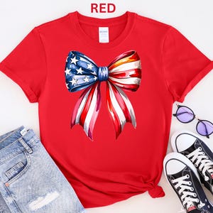 May include: A red t-shirt featuring a watercolor bow design in the colors of the American flag. The bow has blue stars and red and white stripes. The word "RED" is printed at the top. The shirt is paired with blue jeans and black sneakers.
