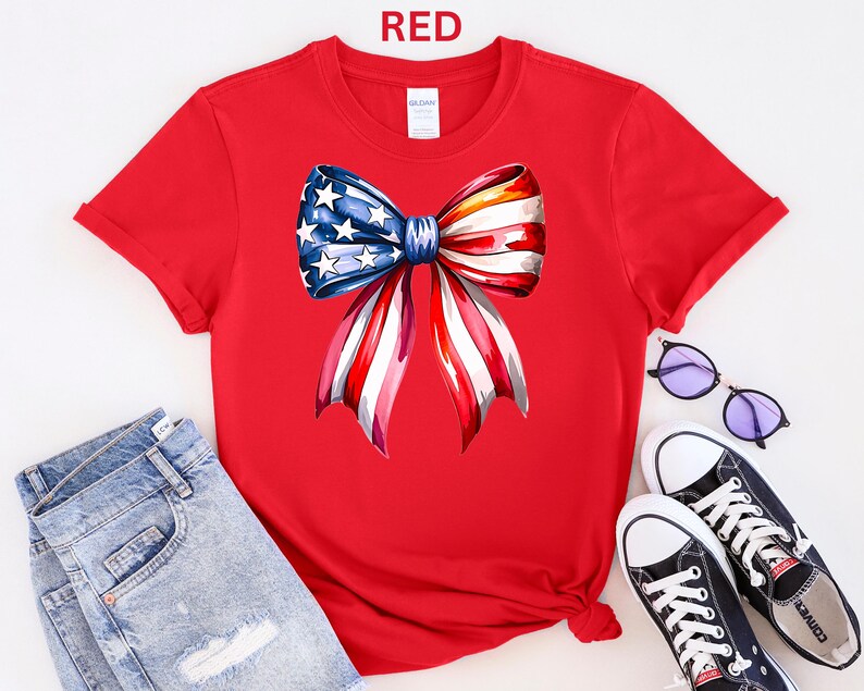 May include: A red t-shirt featuring a watercolor bow design in the colors of the American flag. The bow has blue stars and red and white stripes. The word "RED" is printed at the top. The shirt is paired with blue jeans and black sneakers.