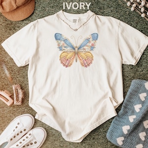 May include: Ivory-colored t-shirt featuring a colorful butterfly design. The butterfly has patchwork-style wings in shades of blue, yellow, pink, and floral patterns. The shirt has a crew neck and short sleeves. A necklace is visible.