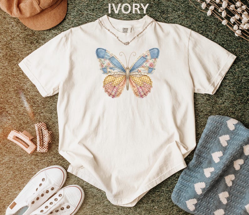 May include: Ivory-colored t-shirt featuring a colorful butterfly design. The butterfly has patchwork-style wings in shades of blue, yellow, pink, and floral patterns. The shirt has a crew neck and short sleeves. A necklace is visible.