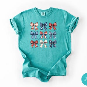 May include: A seafoam green short-sleeve t-shirt featuring a grid of nine decorative bows in various red, white, and blue patterns, including stripes, polka dots, and stars, arranged in three rows on the center of the chest area.