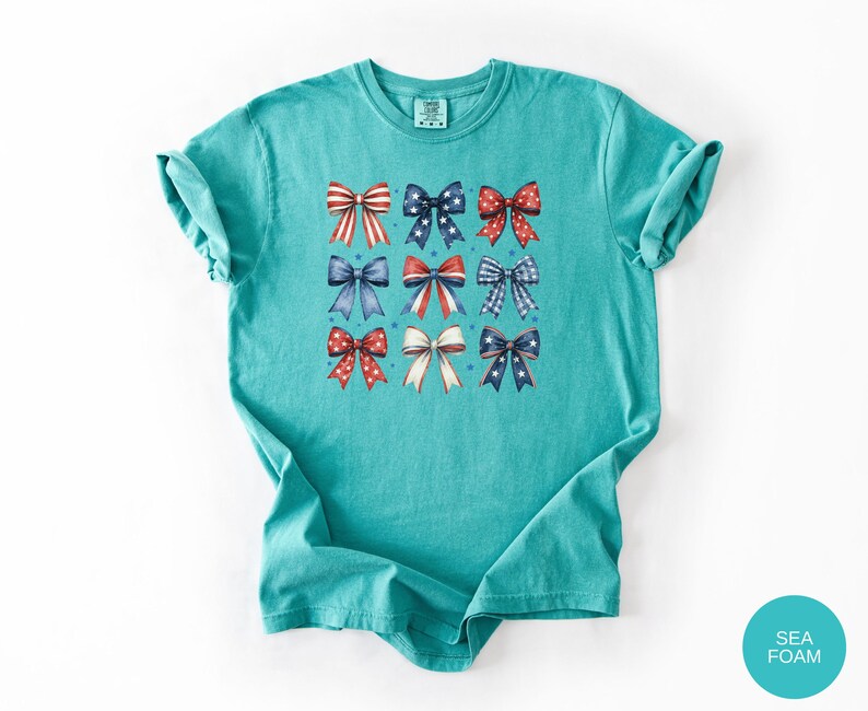 May include: A seafoam green short-sleeve t-shirt featuring a grid of nine decorative bows in various red, white, and blue patterns, including stripes, polka dots, and stars, arranged in three rows on the center of the chest area.