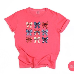 May include: A watermelon-colored short-sleeve t-shirt featuring a central graphic print of nine decorative bows arranged in a grid. The bows display various red, white, and blue patterns, including stars, stripes, and polka dots, with small blue dots.