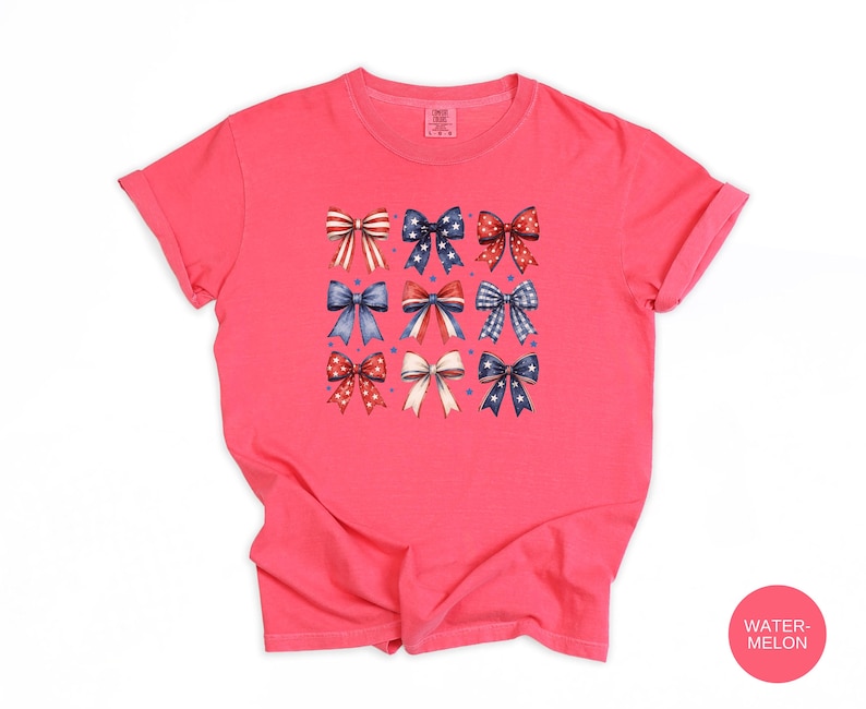 May include: A watermelon-colored short-sleeve t-shirt featuring a central graphic print of nine decorative bows arranged in a grid. The bows display various red, white, and blue patterns, including stars, stripes, and polka dots, with small blue dots.