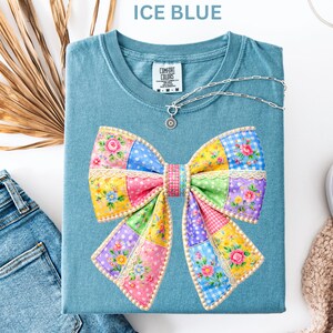 May include: Ice blue t-shirt with a large, colourful bow design. The bow features floral and patchwork patterns in pink, yellow, blue, and green. A silver necklace with a pendant rests on the shirt. The shirt is folded with a pair of jeans and a shoe in the background.