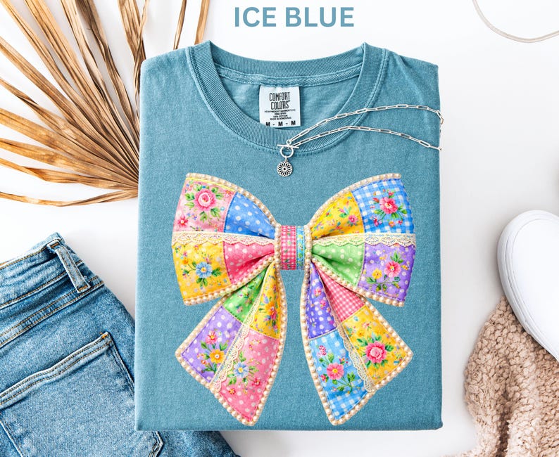 May include: Ice blue t-shirt with a large, colourful bow design. The bow features floral and patchwork patterns in pink, yellow, blue, and green. A silver necklace with a pendant rests on the shirt. The shirt is folded with a pair of jeans and a shoe in the background.