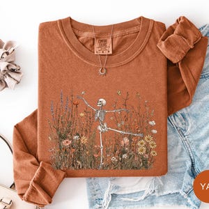 Puede incluir: Una camiseta de manga larga y cuello redondo de color &oacute;xido con un gr&aacute;fico de un esqueleto bailando en un campo de flores silvestres. La prenda tiene una etiqueta de la marca Comfort Colors en el cuello. Un collar dorado con colgante de sol.