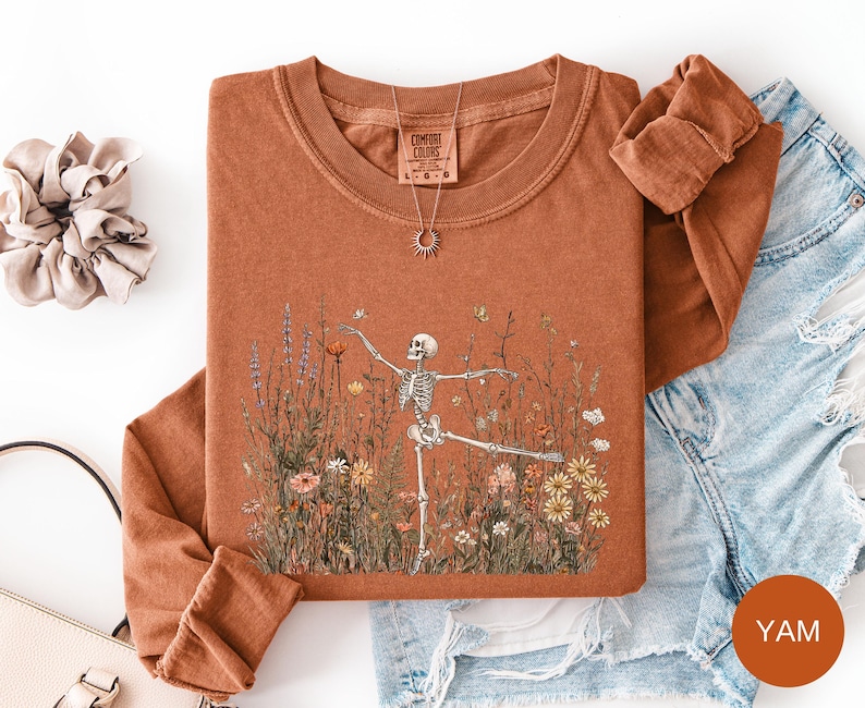 Puede incluir: Una camiseta de manga larga y cuello redondo de color &oacute;xido con un gr&aacute;fico de un esqueleto bailando en un campo de flores silvestres. La prenda tiene una etiqueta de la marca Comfort Colors en el cuello. Un collar dorado con colgante de sol.