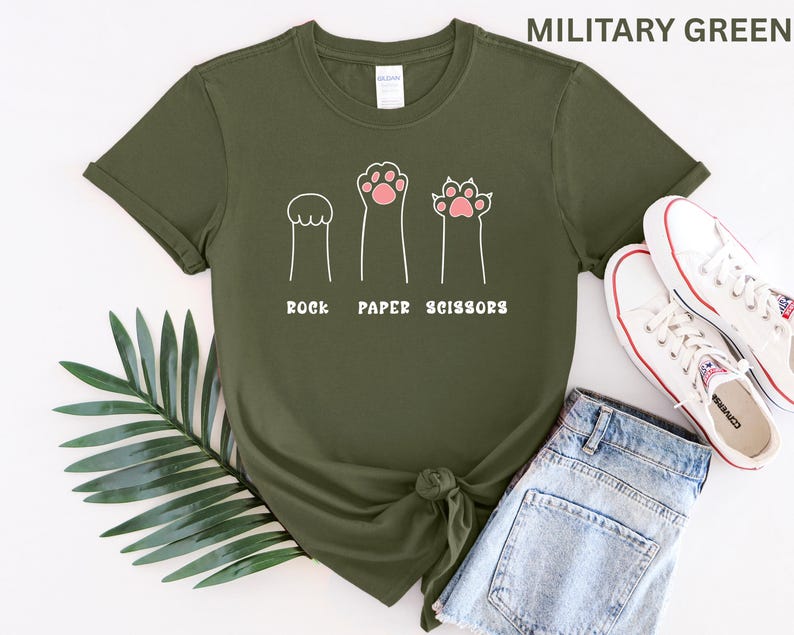 Puede incluir: Camiseta verde militar con un dise&ntilde;o l&uacute;dico de "Piedra, Papel o Tijeras" con ilustraciones de patas de dibujos animados. La camiseta se combina con pantalones cortos de mezclilla y zapatillas blancas con detalles rojos.