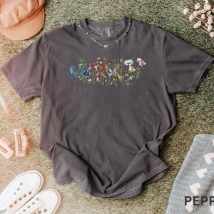 May include: A dark gray t-shirt with a floral design across the chest. The design features a variety of colorful flowers. The shirt is paired with white sneakers, a pink hat, and a decorative necklace. The word "PEPPER" is visible in the bottom right corner.