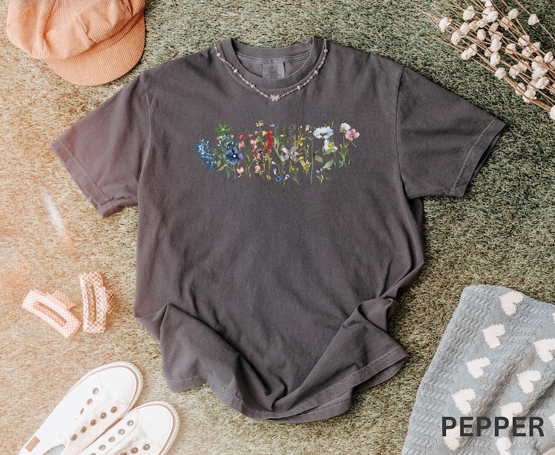 May include: A dark gray t-shirt with a floral design across the chest. The design features a variety of colorful flowers. The shirt is paired with white sneakers, a pink hat, and a decorative necklace. The word "PEPPER" is visible in the bottom right corner.