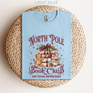 A light blue t-shirt with a Christmas-themed design, including a gingerbread man, presents, and a hot chocolate mug. The text on the t-shirt reads "North Pole Book Club" and "Hot Cocoa Served Daily".