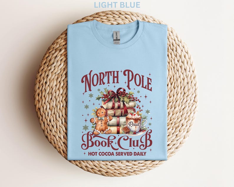 A light blue t-shirt with a Christmas-themed design, including a gingerbread man, presents, and a hot chocolate mug. The text on the t-shirt reads "North Pole Book Club" and "Hot Cocoa Served Daily".