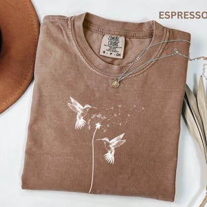 a brown t-shirt with a white bird design, a brown hat, a necklace, and some dried leaves, all arranged on a white background.