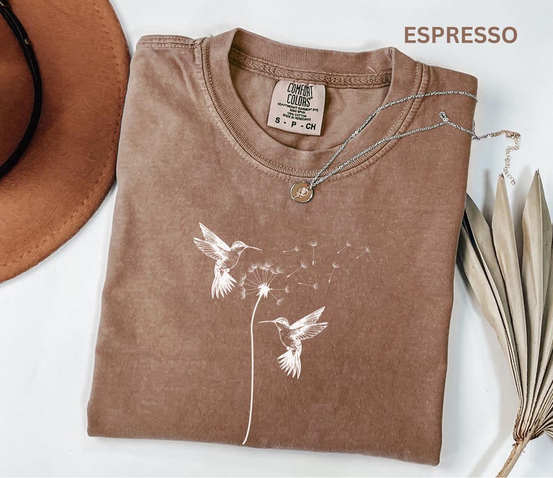 a brown t-shirt with a white bird design, a brown hat, a necklace, and some dried leaves, all arranged on a white background.