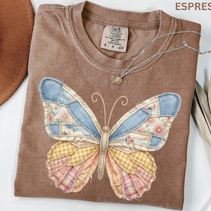 May include: Espresso-colored Comfort Colors t-shirt with a patchwork butterfly design. The butterfly wings feature denim, floral, and plaid patterns in shades of blue, pink, and yellow. A silver necklace with a small pendant rests on the shirt.