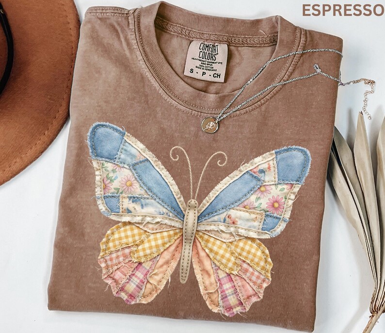 May include: Espresso-colored Comfort Colors t-shirt with a patchwork butterfly design. The butterfly wings feature denim, floral, and plaid patterns in shades of blue, pink, and yellow. A silver necklace with a small pendant rests on the shirt.