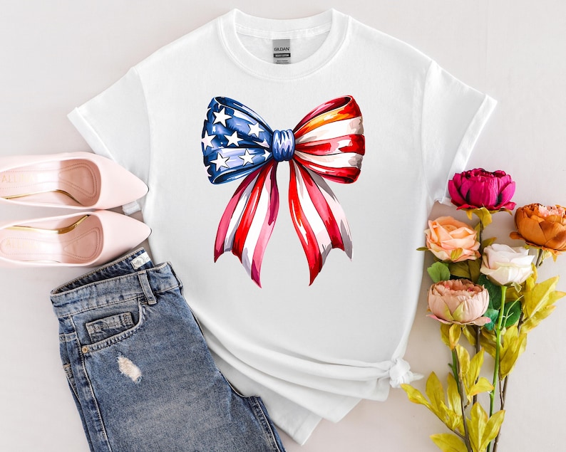 May include: White t-shirt featuring a watercolor bow design in red, white, and blue, with stars on the blue section. The shirt is paired with blue jeans and pink shoes, with a bouquet of flowers.
