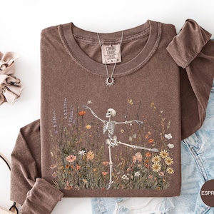 Puede incluir: Una camiseta de manga larga marr&oacute;n con un gr&aacute;fico de un esqueleto bailando en medio de un campo de flores silvestres coloridas. La camiseta tiene cuello redondo y una etiqueta de la marca Comfort Colors. Se muestra un collar con dije de sol.