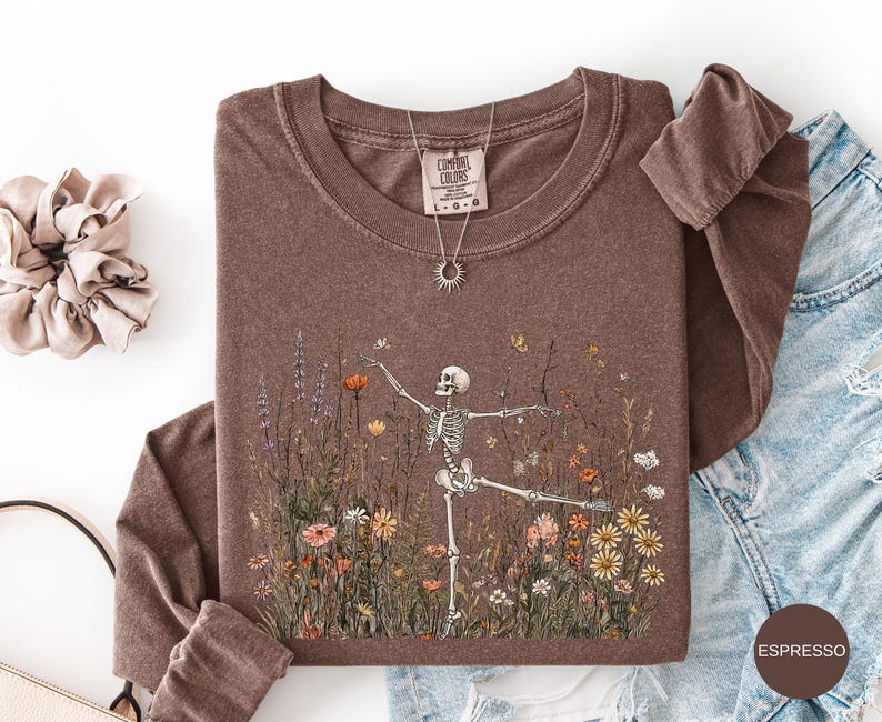 Puede incluir: Una camiseta de manga larga marr&oacute;n con un gr&aacute;fico de un esqueleto bailando en medio de un campo de flores silvestres coloridas. La camiseta tiene cuello redondo y una etiqueta de la marca Comfort Colors. Se muestra un collar con dije de sol.