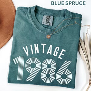 May include: A blue spruce t-shirt with the text "VINTAGE 1986" in white. The shirt has a crew neck and short sleeves. A gold necklace with a pendant is draped on the shirt. A brown hat is visible in the upper left corner.