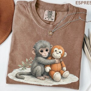 Puede incluir: Camiseta color espresso con un gr&aacute;fico de dos monos abraz&aacute;ndose. Un mono es gris, el otro es naranja. La camiseta tiene cuello redondo y una peque&ntilde;a etiqueta cerca del cuello. Un collar plateado con un peque&ntilde;o colgante est&aacute; drapeado sobre la camiseta.