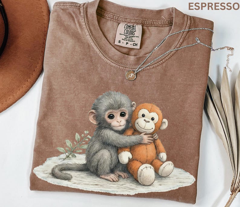 Puede incluir: Camiseta color espresso con un gr&aacute;fico de dos monos abraz&aacute;ndose. Un mono es gris, el otro es naranja. La camiseta tiene cuello redondo y una peque&ntilde;a etiqueta cerca del cuello. Un collar plateado con un peque&ntilde;o colgante est&aacute; drapeado sobre la camiseta.