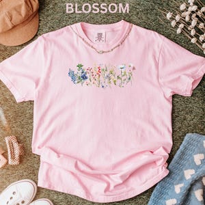 May include: A light pink t-shirt with a floral design across the chest. The word "BLOSSOM" is printed at the top of the image. The shirt is made of a soft material and is likely a comfortable fit.