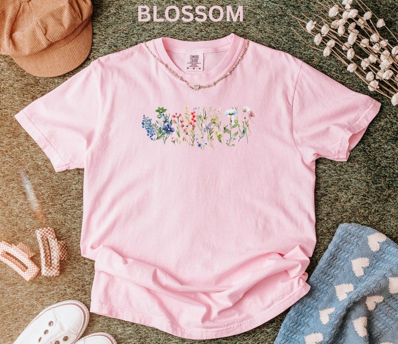 May include: A light pink t-shirt with a floral design across the chest. The word "BLOSSOM" is printed at the top of the image. The shirt is made of a soft material and is likely a comfortable fit.