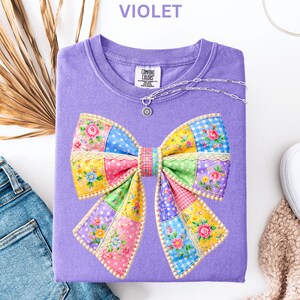 May include: A violet Comfort Colors sweatshirt with a colourful patchwork bow design. The bow features floral, polka dot, and gingham patterns. A silver necklace with a pendant is draped on the shirt. The word "VIOLET" is printed at the top.