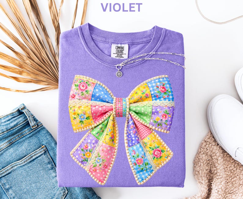 May include: A violet Comfort Colors sweatshirt with a colourful patchwork bow design. The bow features floral, polka dot, and gingham patterns. A silver necklace with a pendant is draped on the shirt. The word "VIOLET" is printed at the top.
