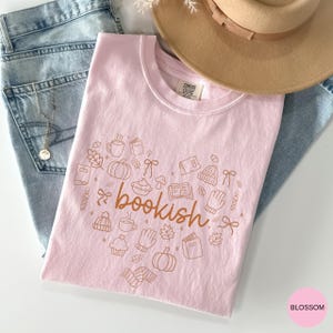 May include: A light pink t-shirt featuring the word bookish in brown cursive text surrounded by brown line art illustrations of pumpkins, books, mugs, leaves, bows, mittens, and beanies. The shirt has a Comfort Colors label at the neck.