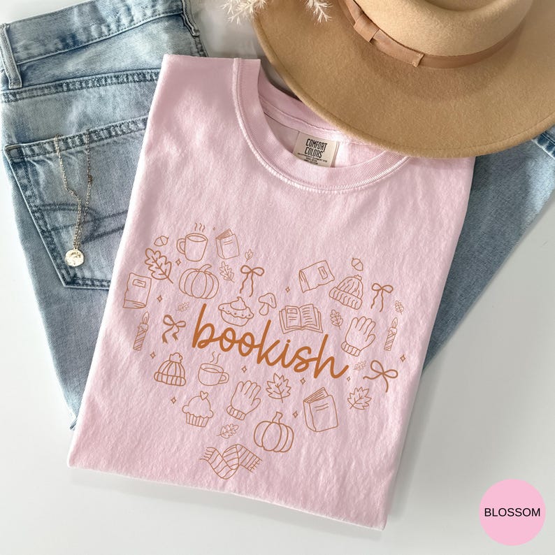 May include: A light pink t-shirt featuring the word bookish in brown cursive text surrounded by brown line art illustrations of pumpkins, books, mugs, leaves, bows, mittens, and beanies. The shirt has a Comfort Colors label at the neck.