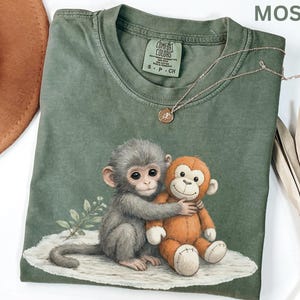 May include: Moss green t-shirt featuring a graphic of a gray monkey hugging an orange stuffed monkey. The shirt has a crew neck and short sleeves. A delicate silver necklace with a small pendant rests on the shirt. The tag reads "COMFORT COLORS".