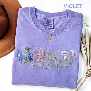 May include: A violet Comfort Colors t-shirt with a floral design across the chest. The design features a variety of colorful wildflowers. A gold necklace with a pendant rests on the shirt. A brown hat and dried plant are in the background.