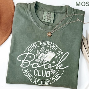 May include: A moss green t-shirt with the text "WHAT HAPPENS AT BOOK CLUB STAYS AT BOOK CLUB" in a white circular design. A gold necklace with a pendant rests on the shirt. The shirt is folded and placed on a white surface.