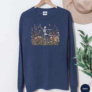 Puede incluir: Una camiseta azul marino de manga larga con un estampado gr&aacute;fico de un esqueleto bailando en un campo de flores silvestres coloridas, incluyendo margaritas, amapolas y varias hierbas. La camiseta tiene cuello redondo y est&aacute; en una percha.