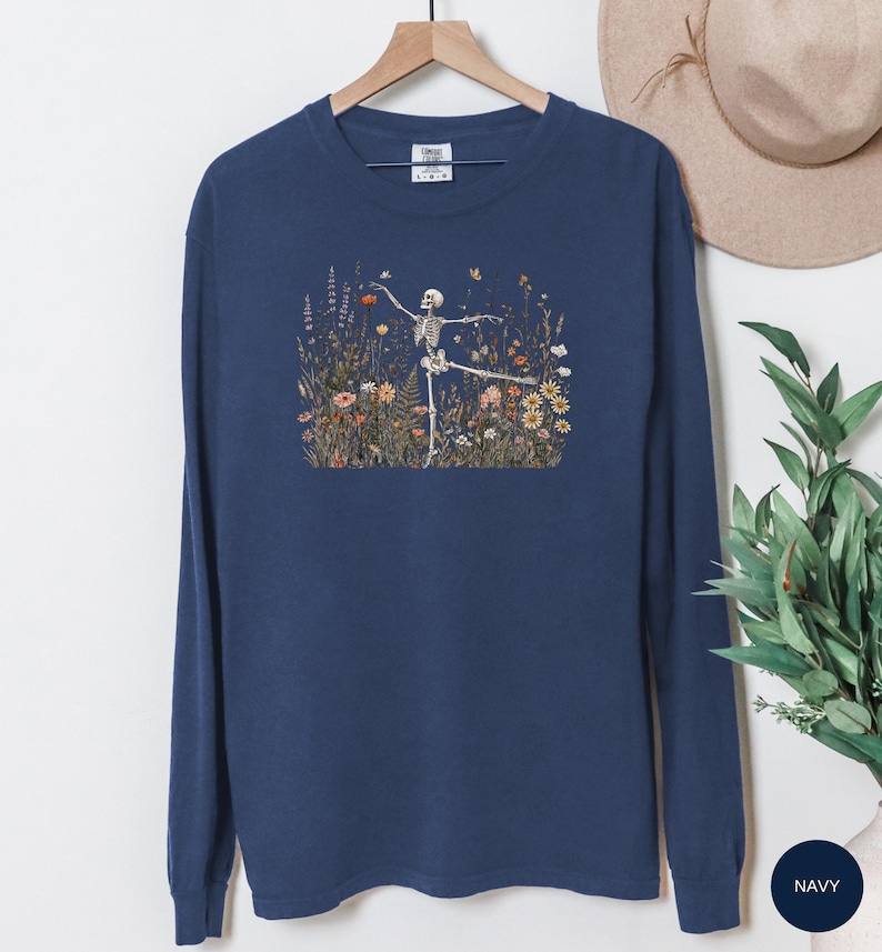 Puede incluir: Una camiseta azul marino de manga larga con un estampado gr&aacute;fico de un esqueleto bailando en un campo de flores silvestres coloridas, incluyendo margaritas, amapolas y varias hierbas. La camiseta tiene cuello redondo y est&aacute; en una percha.
