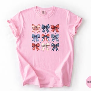 May include: A light pink short-sleeved t-shirt featuring a central graphic print of nine decorative bows arranged in a grid. The bows display various red, white, and blue patterns including stripes, polka dots, and stars, accented by small blue stars.